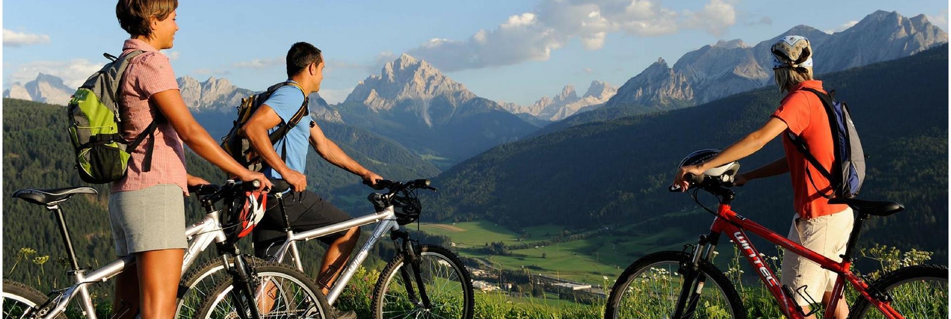 Tre ciclisti con zaini osservano una valle verde e le Dolomiti da un punto panoramico.