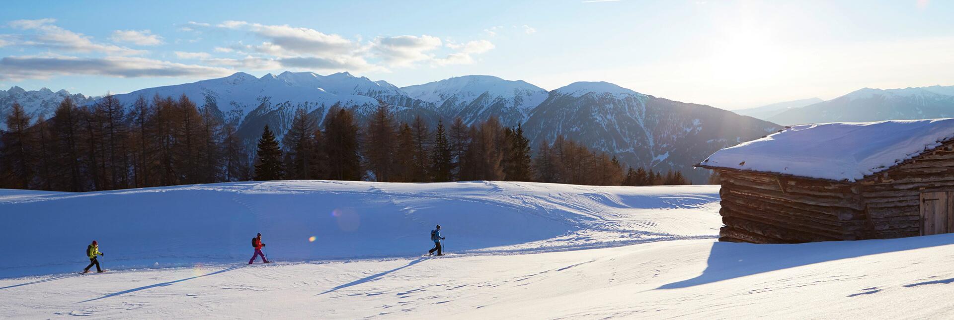 Three people snowshoeing through a snowy winter landscape with a cabin and mountain backdrop.